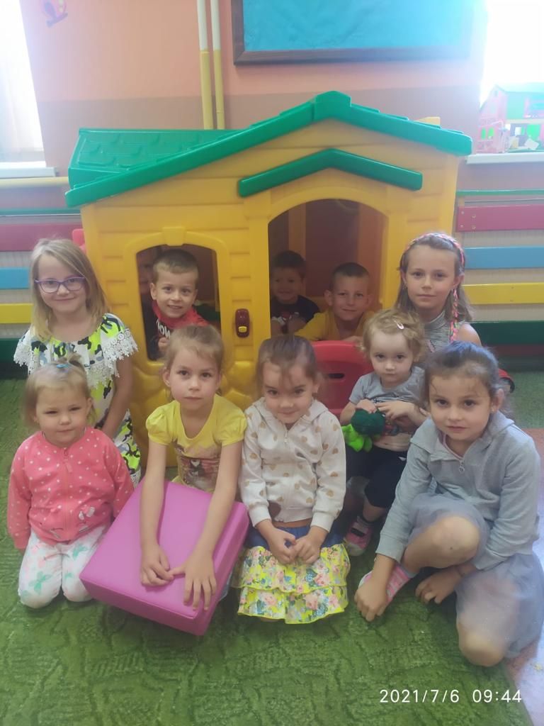 Dzieci przedszkole siedzą na dywanie w sali na tle plastikowego domku do zabawy, niektóre w rękach trzymają siedziska do siedzenia